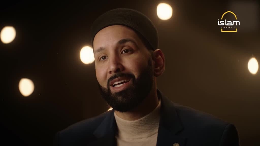 A man with a dark beard and cap speaks, his mouth moving as if he's mid-sentence. Behind him, soft, round lights glow against a dark backdrop, and the Islam Channel logo is visible.
