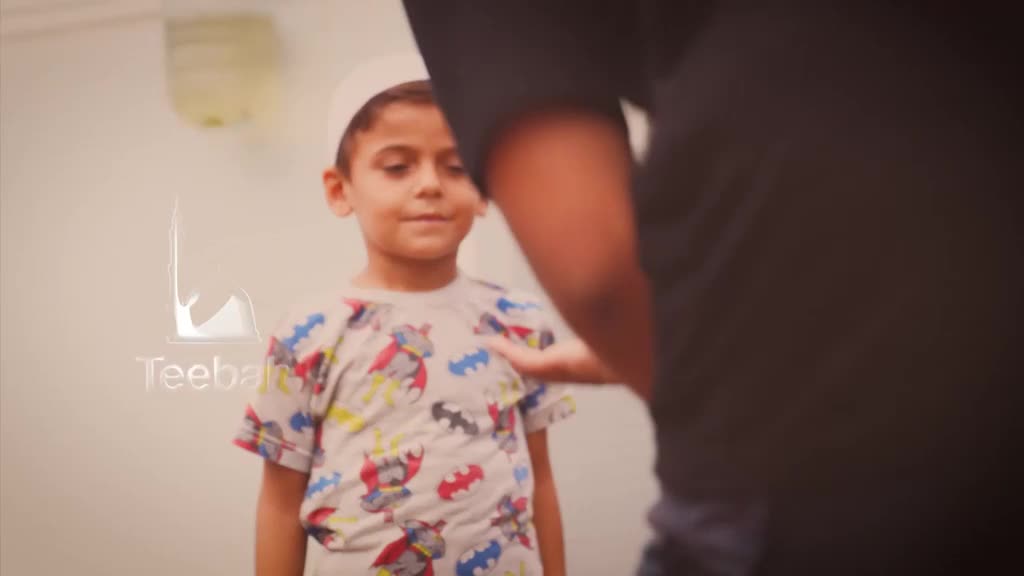A young boy wearing a Batman t-shirt and a white cap stands facing the camera. Someone's hand rests gently on his chest, likely adjusting his position.
