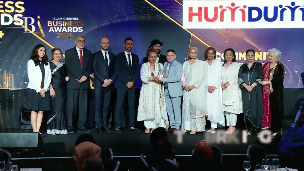 A group of people, some in suits and others in traditional attire, are on stage, holding a trophy. The Islam Channel Business Awards logo is visible in the background.
