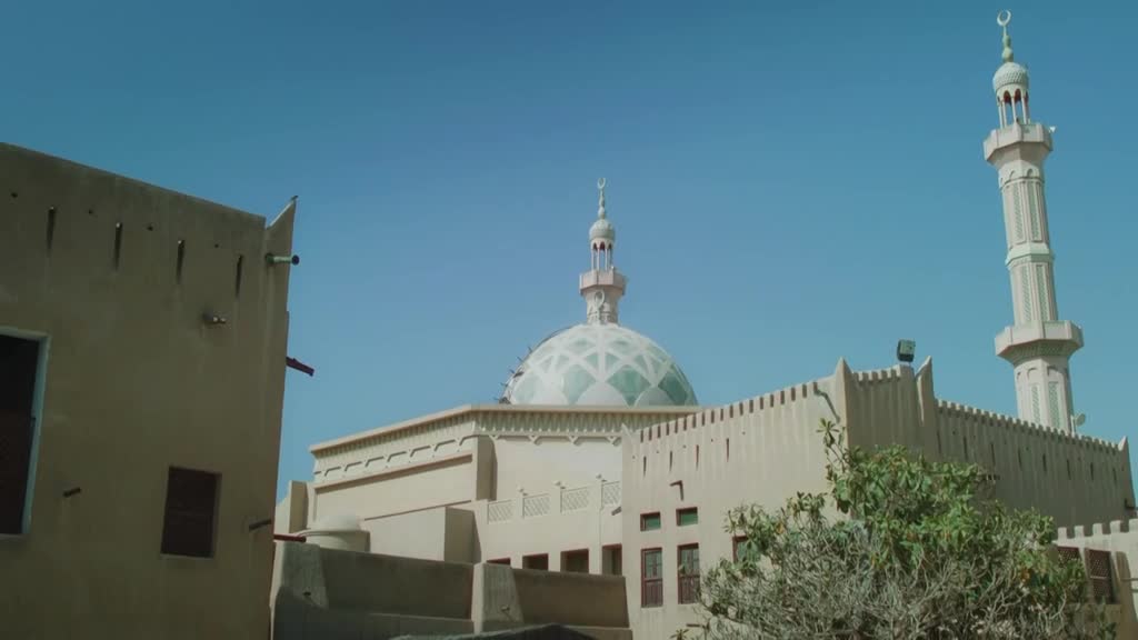 A mosque with a patterned dome and a tall minaret rises against a clear blue sky.  The building's beige walls are punctuated by small windows and a crenellated roofline.
