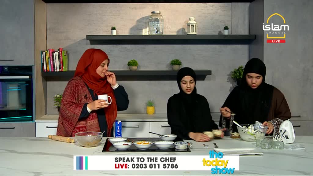 A woman in a red headscarf sips from a mug, while two others, in black, prepare food at a counter. One stirs a bowl with a spoon, and the other arranges something in small glass dishes.

