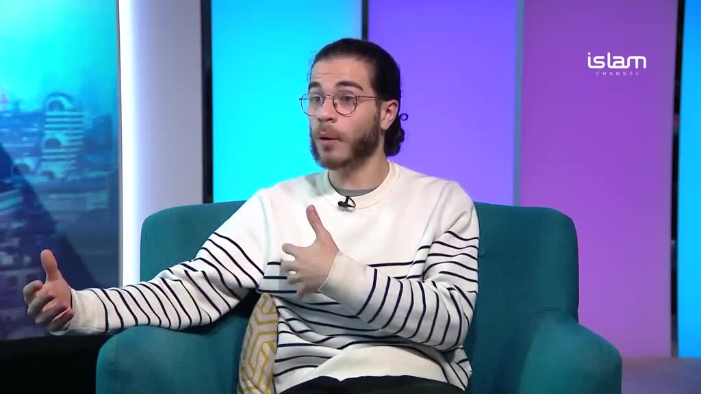 A man in a striped sweater gestures with his hands while speaking, seated on a teal couch. Behind him, the logo for Islam Channel is visible against a backdrop of purple and blue.
A man in a striped sweater gestures with his hands while speaking, seated on a teal couch. Behind him, the logo for Islam Channel is visible against a backdrop of purple and blue.