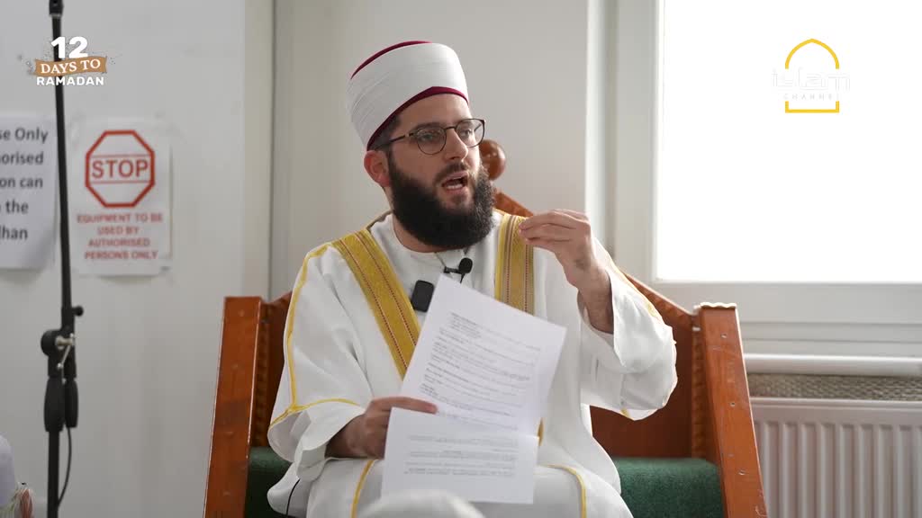 A man in a white robe and turban speaks, holding papers. He gestures with his right hand as he addresses an unseen audience.