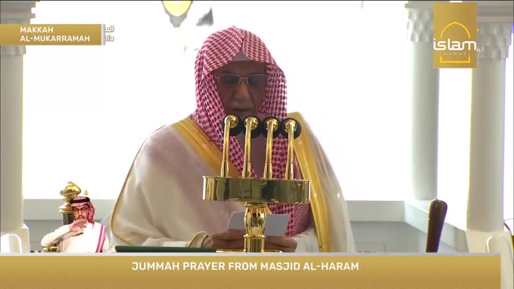 An imam, wearing a red and white checkered ghutra and a white thobe, stands before a golden microphone stand, reading from papers. The Islam Channel logo is visible in the upper right corner, with "JUMMAH PRAYER FROM MASJID AL-HARAM" displayed at the bottom.