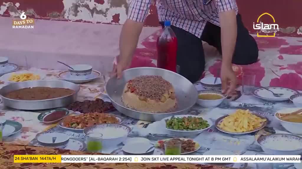 A large, round dish of food sits center, topped with a sprinkle of what looks like ground meat. Plates of various dishes are spread across a patterned cloth, ready for a meal.