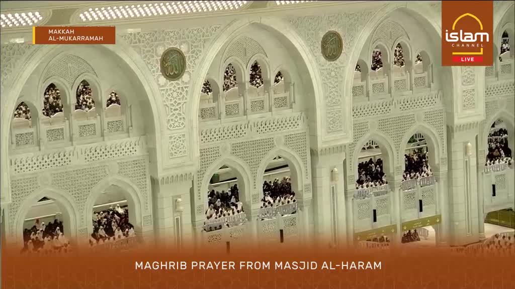 People are gathered in the upper levels of the Masjid al-Haram, looking out over the prayer hall. The intricate white marble architecture of the mosque surrounds them.