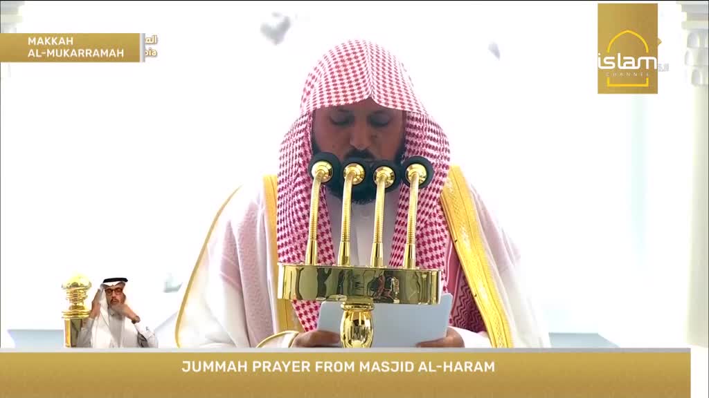 The Imam stands before four microphones, his head bowed as he reads from a text. A smaller screen to the left shows another man in similar attire, also looking down.