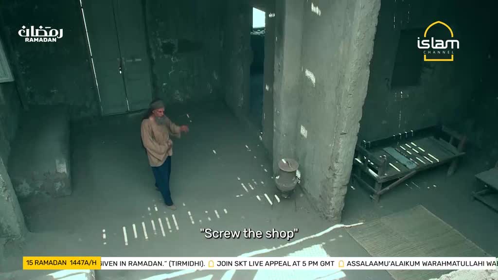 A man with a long beard walks through a dimly lit room, gesturing with his hand. Sunlight streams through gaps in the ceiling, casting sharp patterns on the floor.