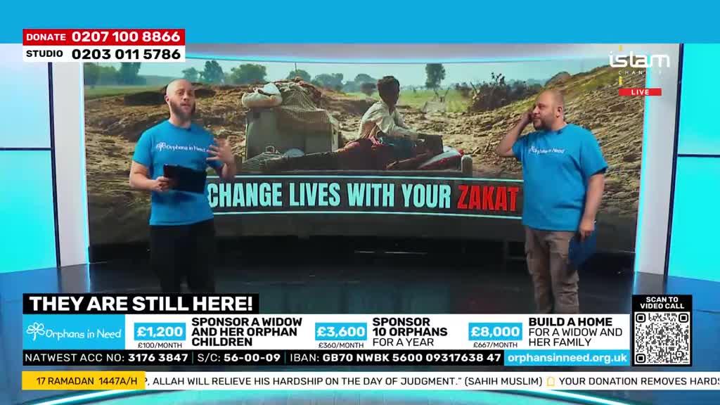 Two men in blue t-shirts stand on a studio set, gesturing towards a large screen displaying a scene of hardship. The screen shows a person lying on a cart in a dusty, arid landscape.