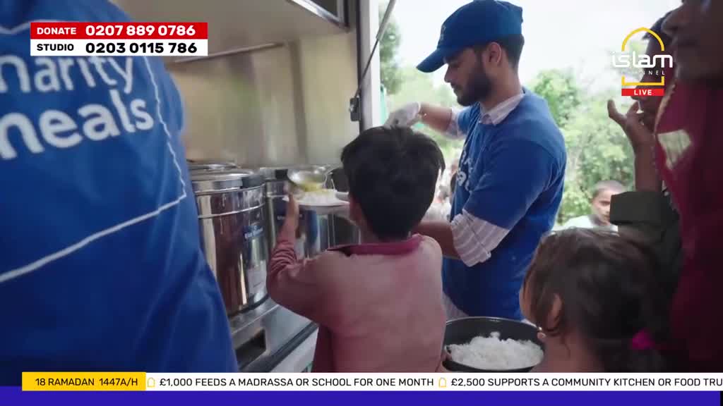 A volunteer serves food from a large metal container to a child. Another volunteer, wearing a blue shirt with "Charity Meals" printed on it, stands nearby.
