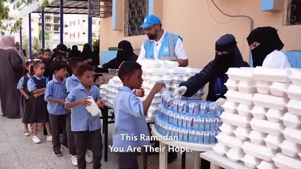 Children in blue uniforms queue patiently as volunteers hand out food parcels. A man in a blue vest and cap supervises the distribution of stacked white containers.