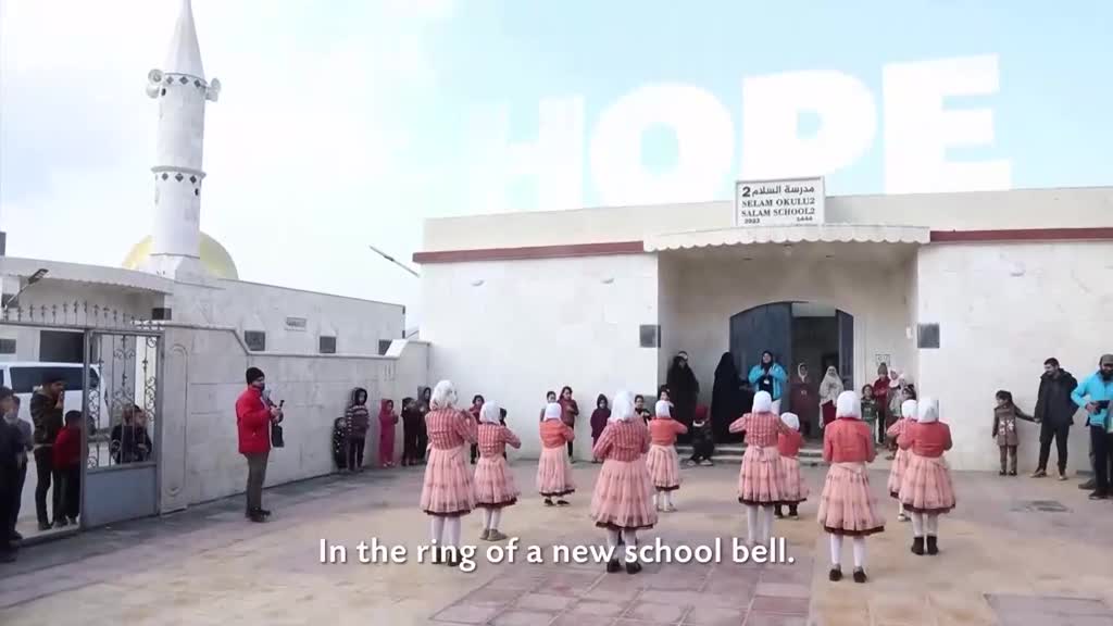 A group of girls in pink dresses and white headscarves are dancing in a courtyard. A man in a red jacket plays a stringed instrument as children watch.