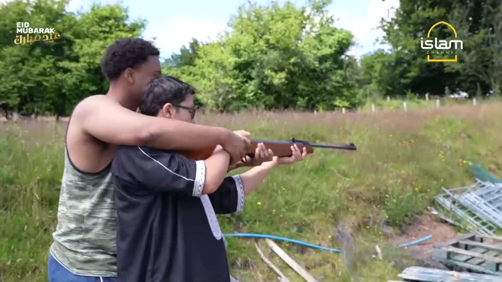 An older boy steadies a younger boy's aim with a rifle in a grassy field. The younger boy, wearing glasses and a black thobe, holds the rifle steady as the older boy's arms wrap around him. An older boy steadies a younger boy's aim with a rifle in a grassy field. The younger boy, wearing glasses and a black thobe, holds the rifle steady as the older boy's arms wrap around him.