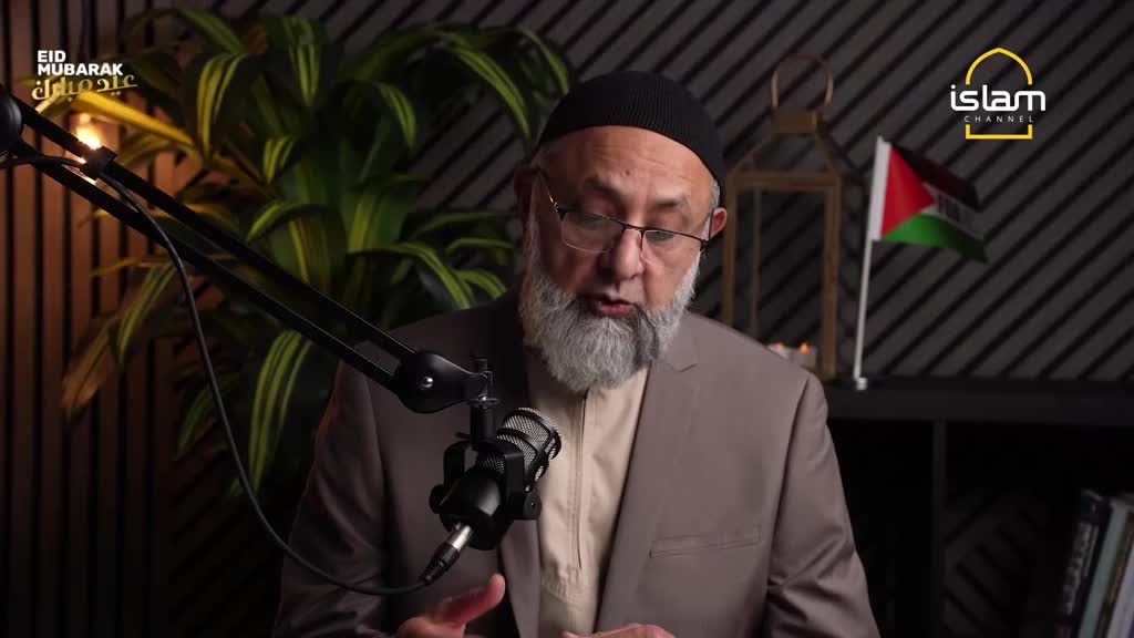 A man with a grey beard and a black cap speaks into a microphone. Behind him, a Palestinian flag hangs beside a decorative lantern. A man with a grey beard and a black cap speaks into a microphone. Behind him, a Palestinian flag hangs beside a decorative lantern.