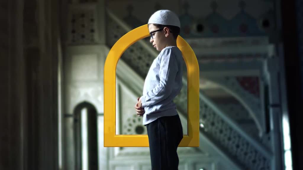 A young boy stands in prayer, his hands clasped before him. He's wearing a white kufi and glasses, framed by a bright yellow archway inside a mosque.