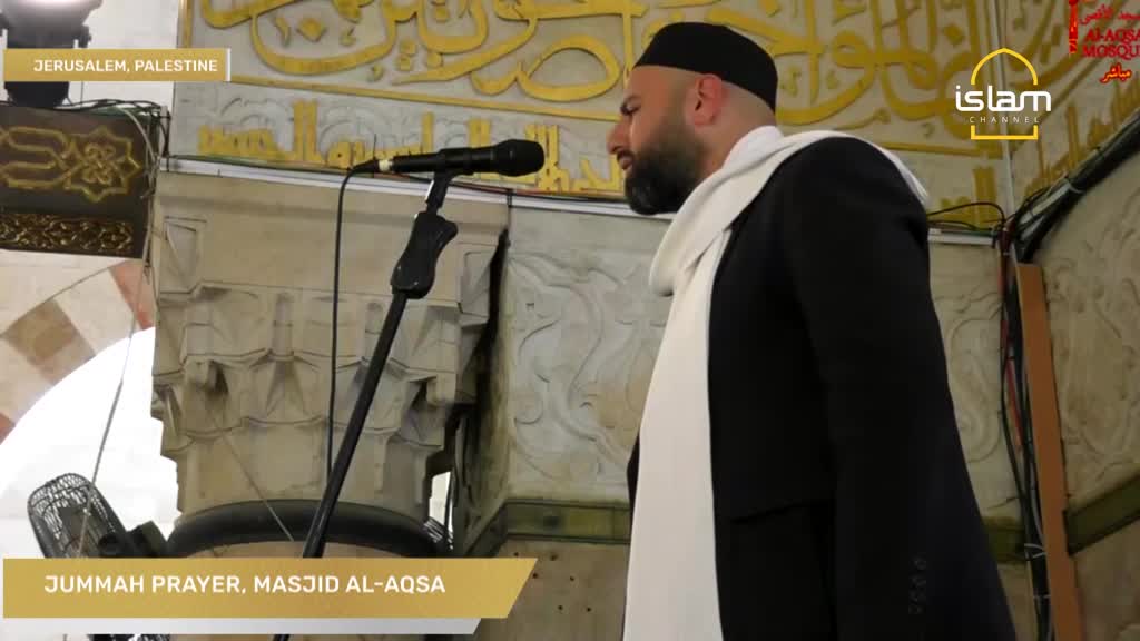 A man in a black suit and white scarf stands before a microphone, his gaze directed upwards. This is the Jumuah prayer at Masjid Al-Aqsa in Jerusalem, Palestine, broadcast on Islam Channel.