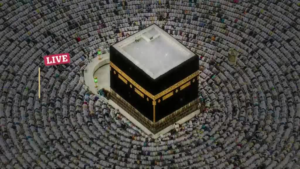 Thousands of people in white robes are prostrating in unison around the Kaaba. A "LIVE" sign hangs in the air, broadcasting this moment to viewers, perhaps in the UK.