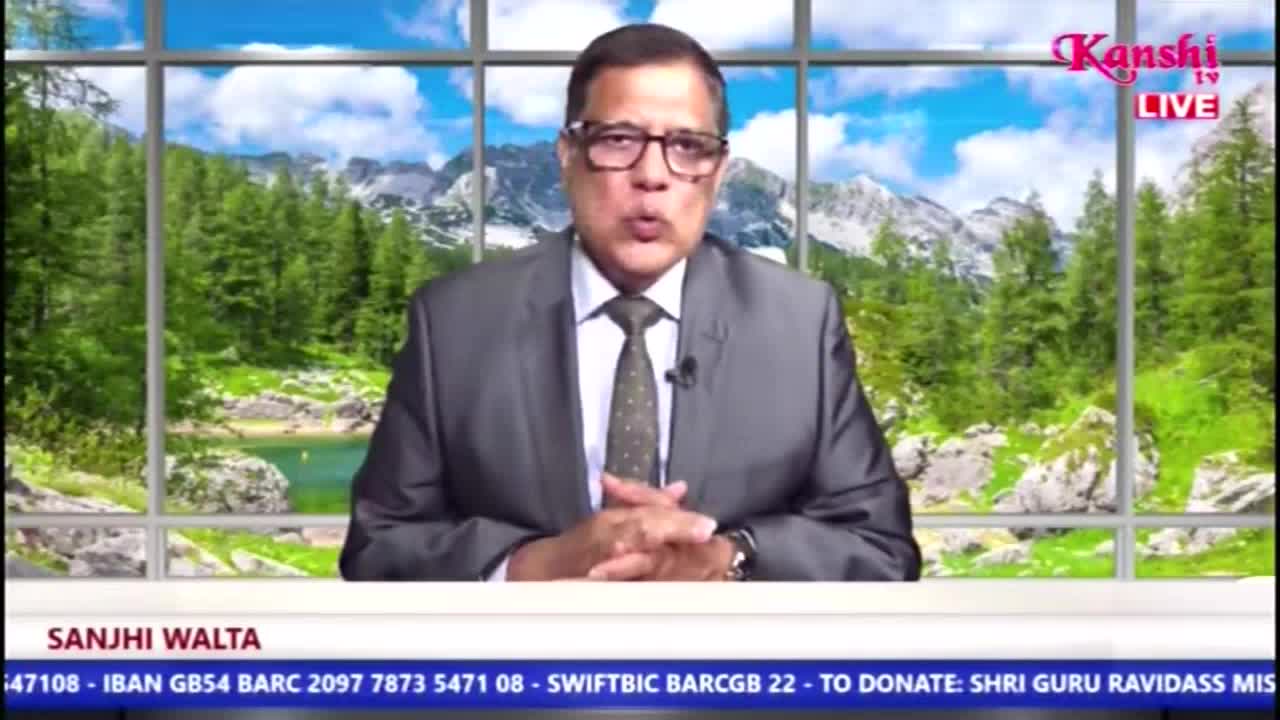 A man in a suit and tie, wearing glasses, is speaking directly to the camera on Kanshi TV, likely a news program from the United Kingdom. Behind him, a backdrop shows a scenic view of mountains and trees.
