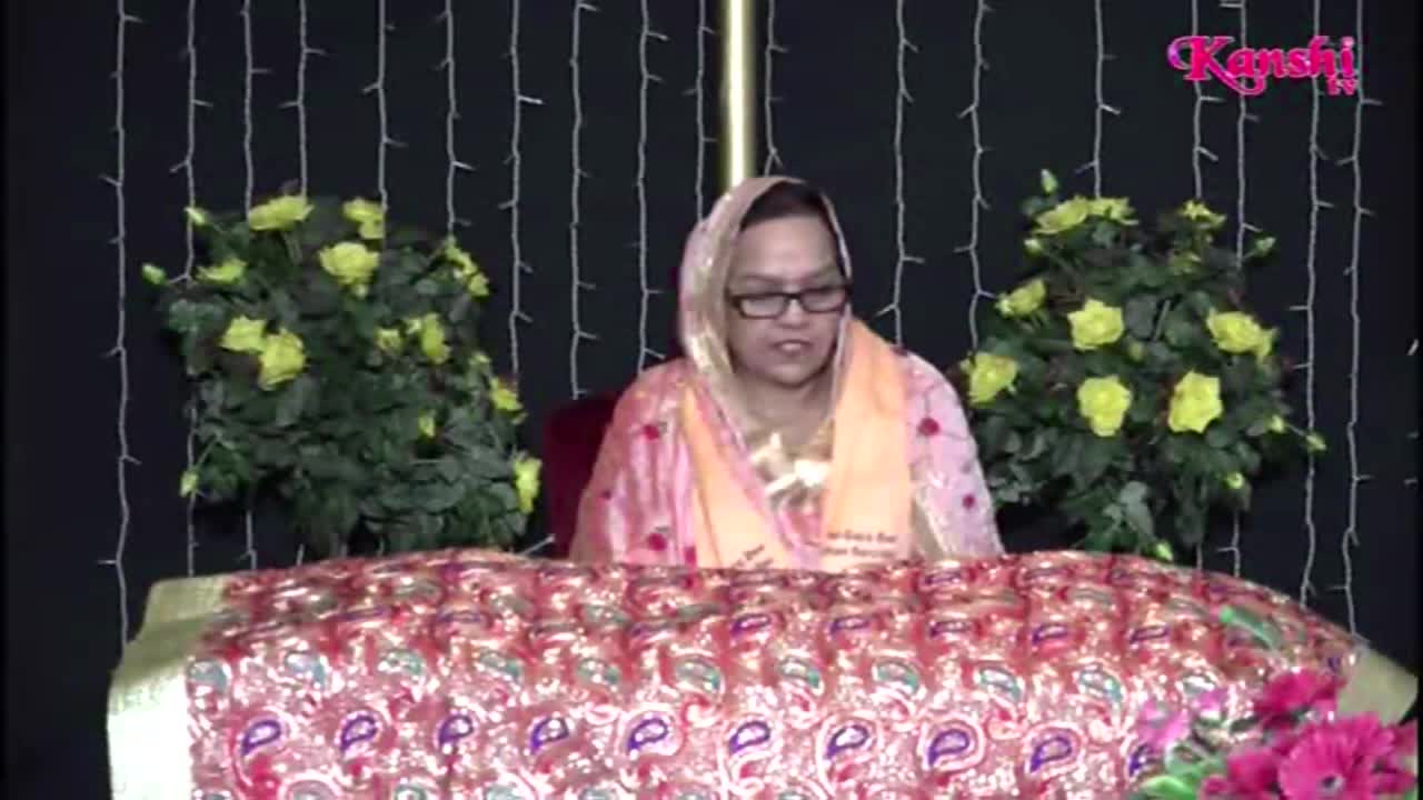 A woman in a headscarf and glasses sits behind a table covered in a patterned cloth, speaking into a microphone. Behind her, on either side, are large arrangements of yellow flowers, and the Kanshi TV logo is visible in the upper right corner.
