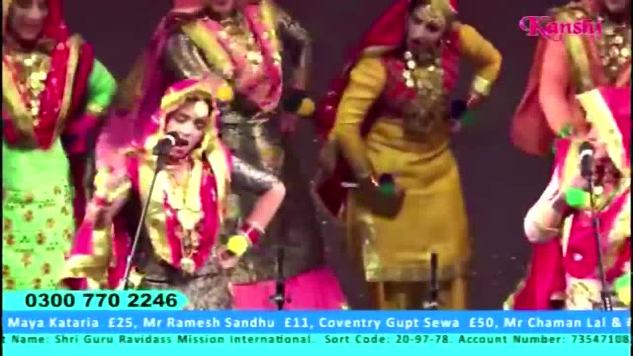 On Kanshi TV, a group of performers in vibrant traditional dress are dancing and singing. The woman in the foreground is holding a microphone, while the others move in unison.
