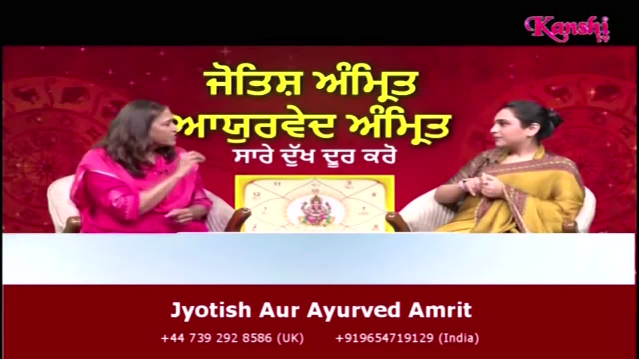 Two women are seated, facing each other on a Kanshi TV set. The woman in pink gestures as she speaks, while the other listens, a gold shawl draped over her shoulders.
