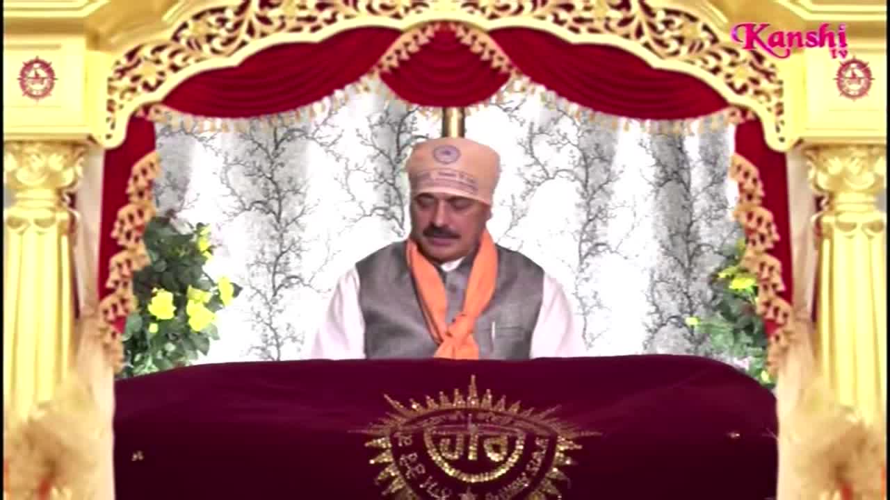 A man in a turban and orange scarf sits behind a decorated table, speaking into a microphone. The Kanshi TV logo is visible in the top right corner.
