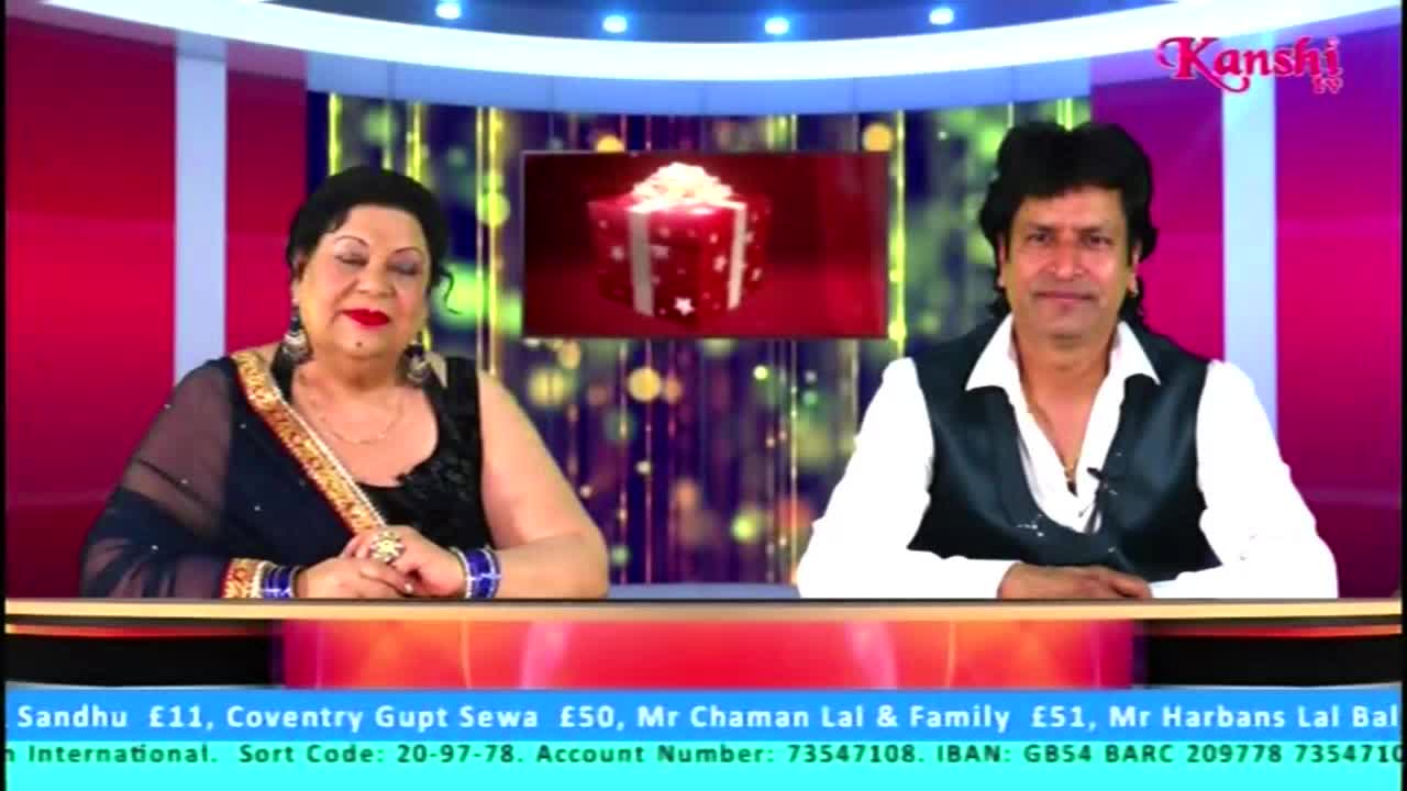 Two people sit behind a desk, the woman in a dark sari and the man in a vest and open shirt. A screen behind them on Kanshi TV shows a gift box animation.
