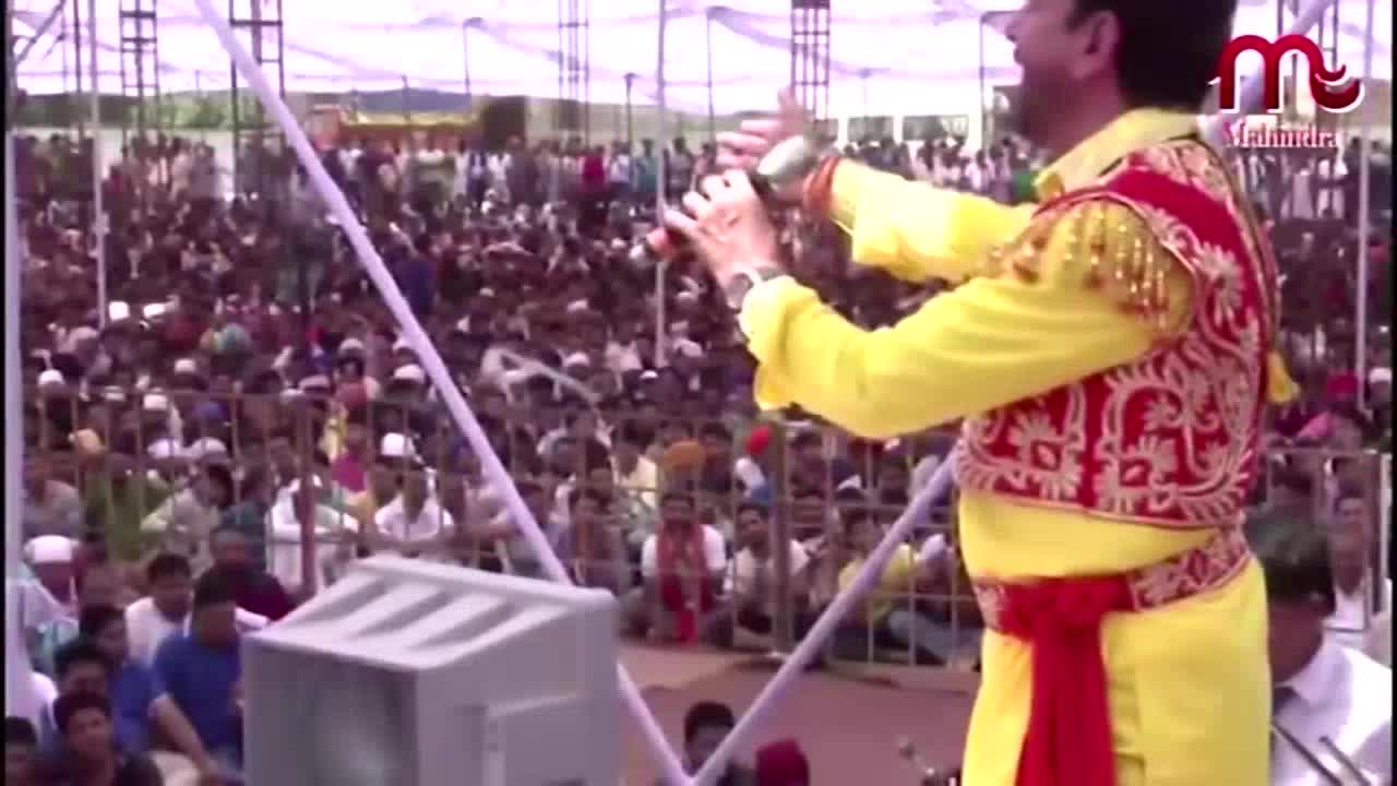 A man in a bright yellow outfit with red embroidery gestures with a microphone towards a large crowd. The Kanshi TV logo is visible in the corner, and the stage is set up outdoors.
