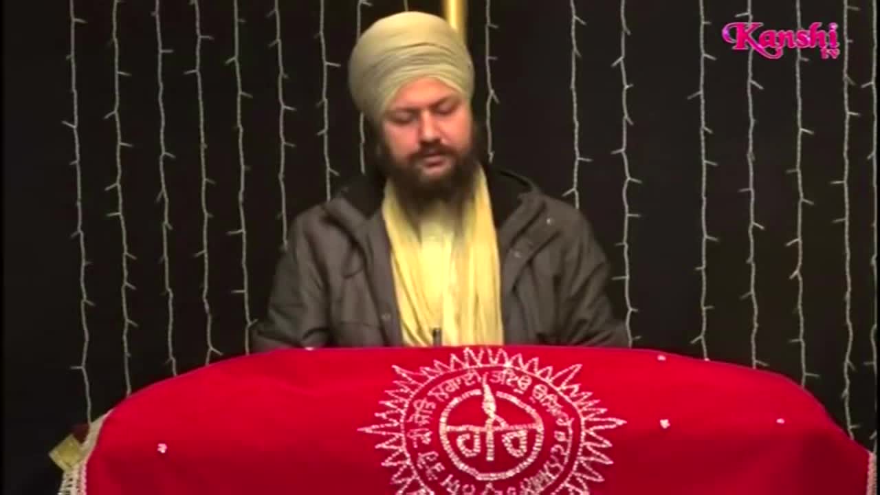 A man in a turban sits behind a red cloth, speaking on Kanshi TV. Behind him, tiny white lights hang against a dark backdrop.
