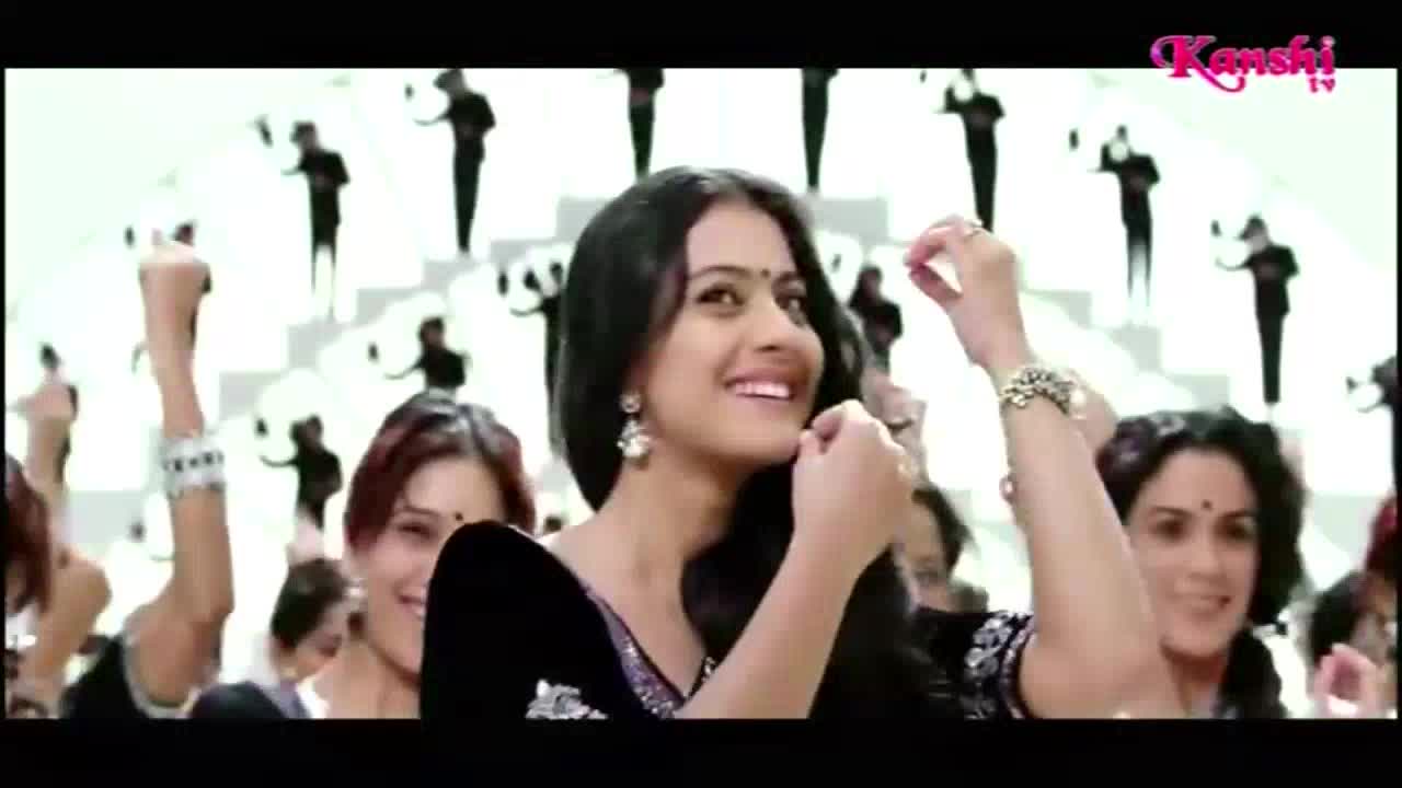 A woman in a black top smiles, her hand touching her hair as she dances. Behind her, a crowd of women and men in silhouette move on a white background, all part of the Kanshi TV production.
