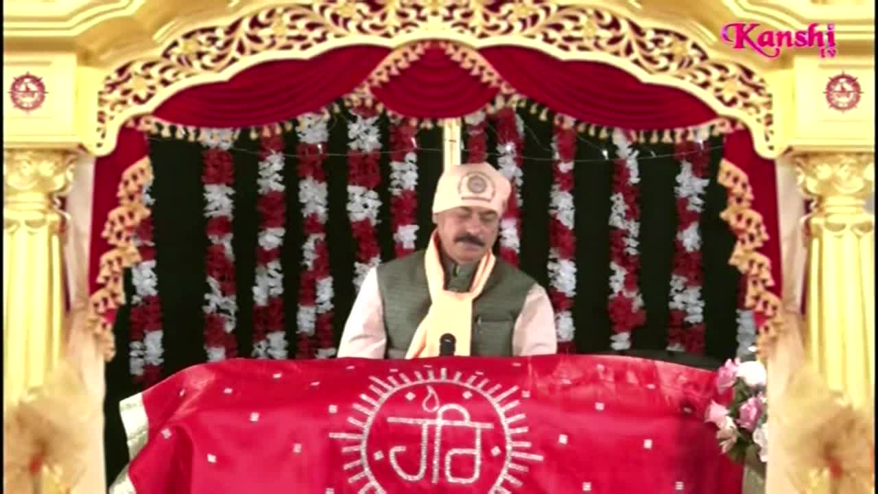 A man in a turban and vest stands behind a red-covered table, speaking into a microphone. Behind him, a backdrop of red and white floral garlands hangs, and the Kanshi TV logo is visible.

