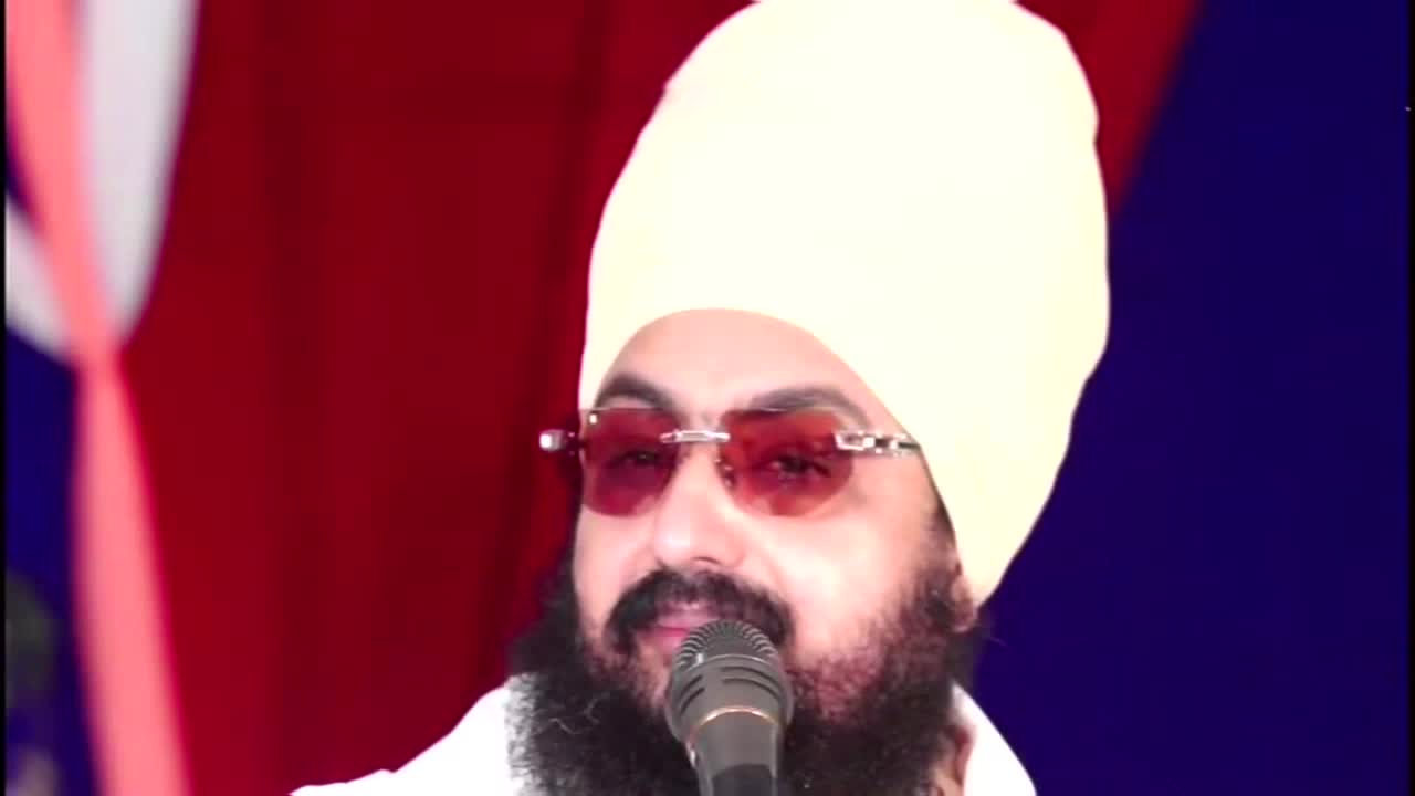 A man in a turban and sunglasses speaks into a microphone, his beard framing his face. Behind him, a red and blue backdrop provides a simple stage setting, likely for a Kanshi TV broadcast from the United Kingdom.
