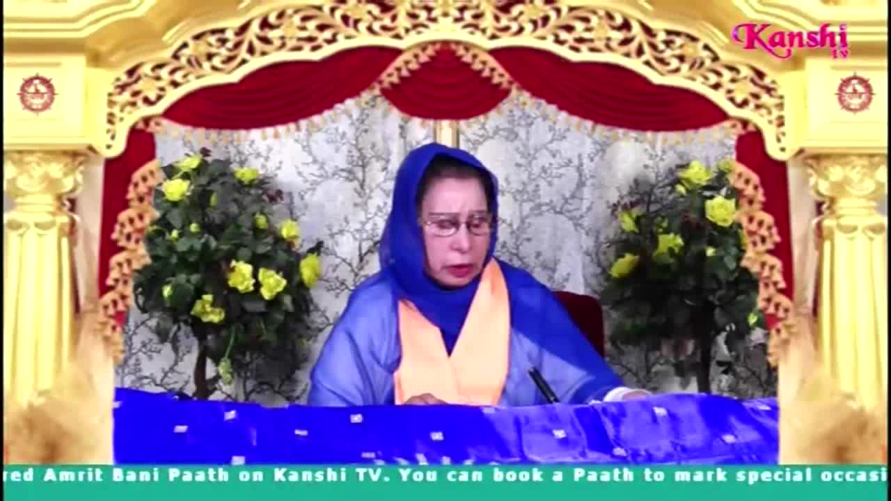 A woman in a blue head covering and glasses speaks, seated at a table draped in blue fabric. The Kanshi TV logo is visible in the upper right corner, and the backdrop is ornate with gold and red accents.
