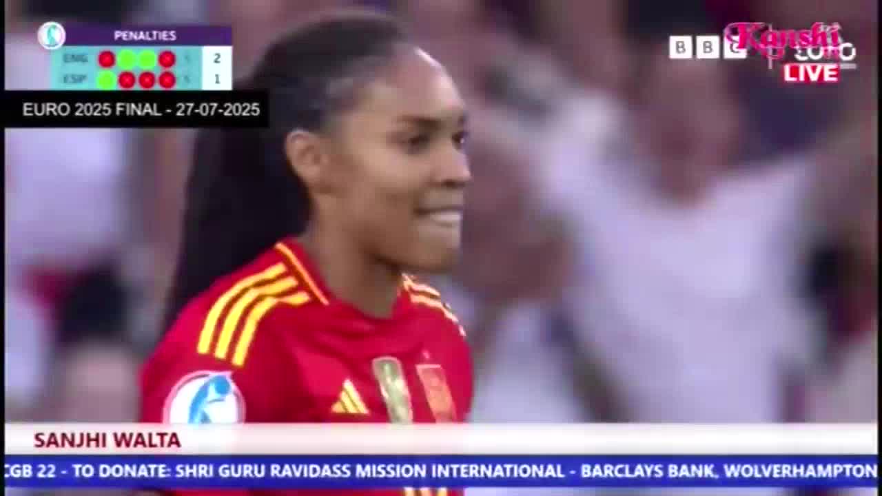 The player in the red and yellow jersey is smiling, looking toward the field. The scoreboard shows the penalties in the Euro 2025 final, broadcast on Kanshi TV in the United Kingdom.
