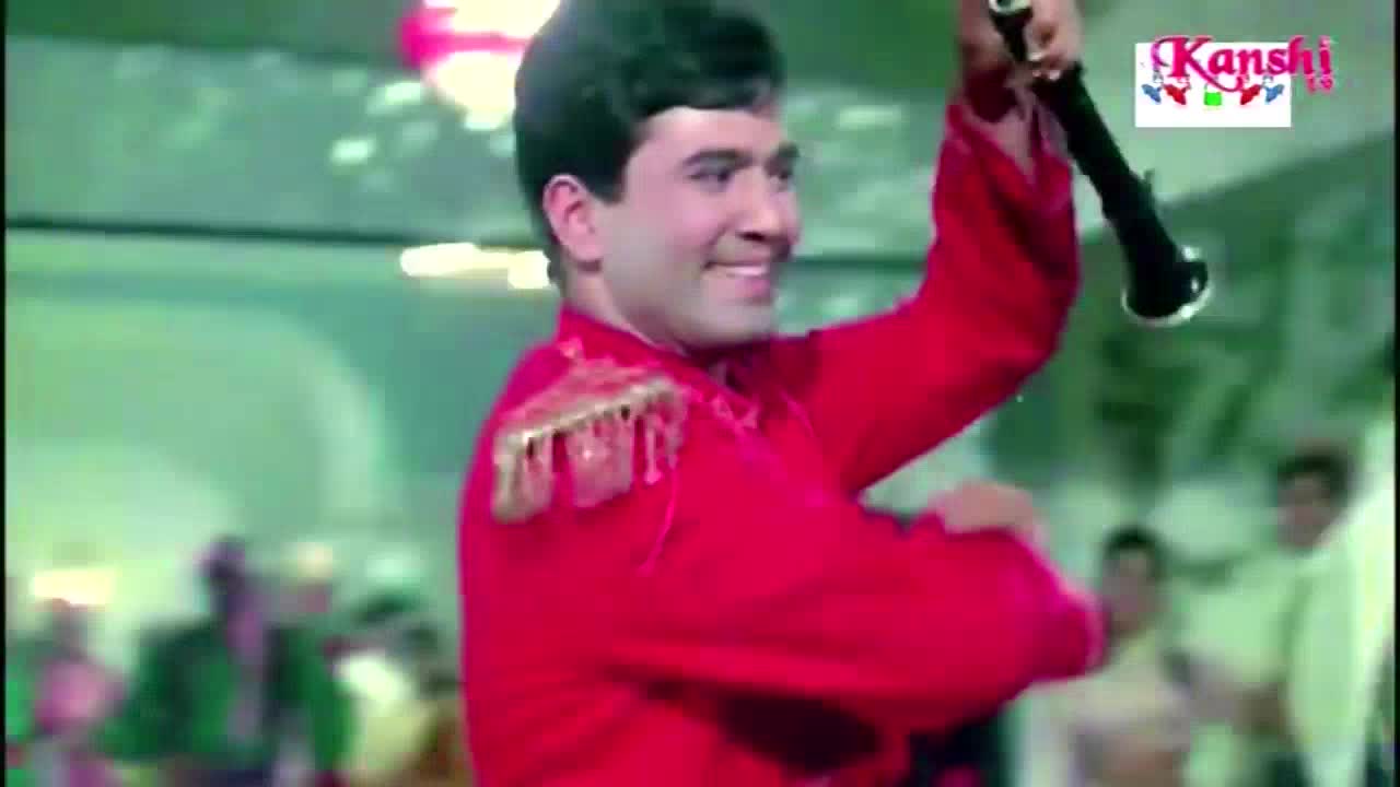 A man in a bright red jacket with gold trim is smiling, holding a black instrument aloft. The Kanshi TV logo is visible in the corner.
