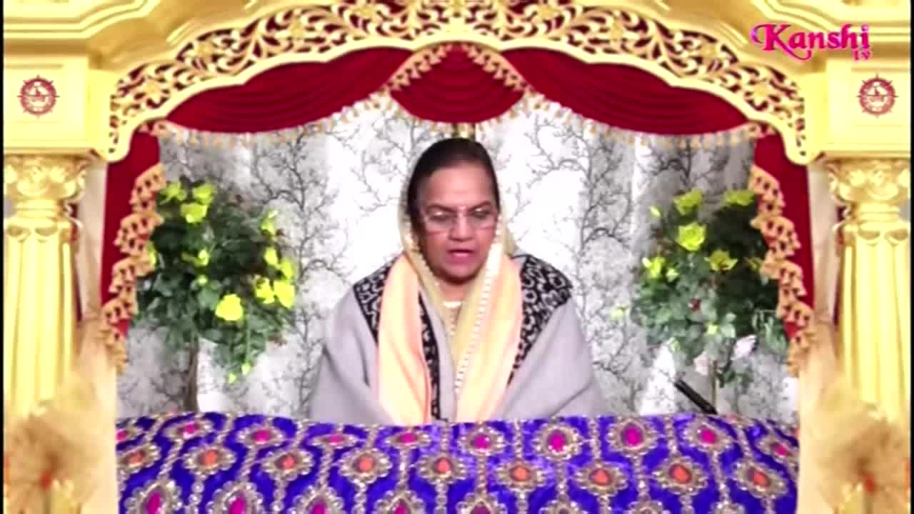 A woman with glasses, wearing a shawl, speaks from behind a decorated table. The Kanshi TV logo is visible in the upper right corner.
