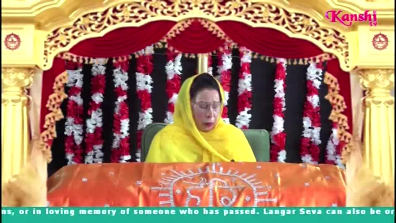 A woman in a yellow head covering is seated behind a table, speaking into a microphone. The Kanshi TV logo is visible in the upper right corner, and the backdrop is decorated with red and white floral arrangements.
