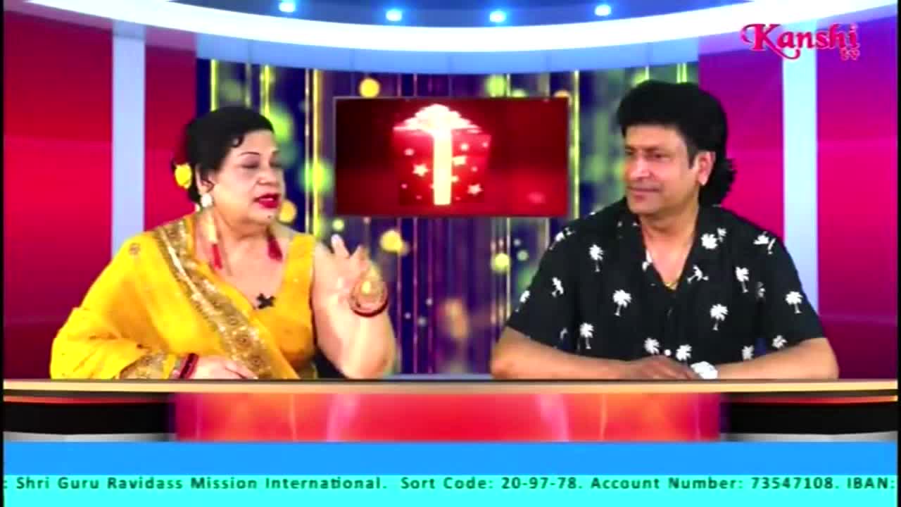 On the Kanshi TV set, a woman in a yellow sari gestures with her right hand while speaking. Beside her, a man in a black shirt with white palm tree patterns listens, seated at a desk.
