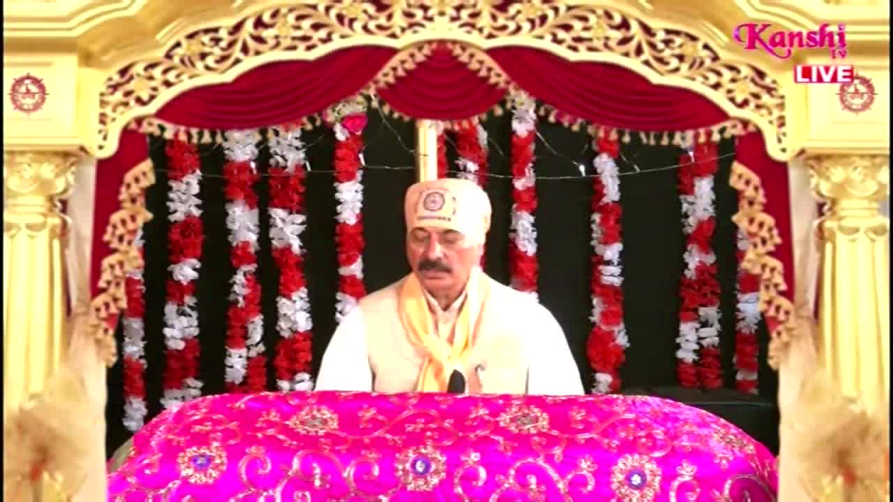A man in a turban sits behind a table covered in a bright pink cloth, speaking into a microphone. Kanshi TV's logo is visible in the corner, and the backdrop is elaborately decorated with red and white floral garlands.
