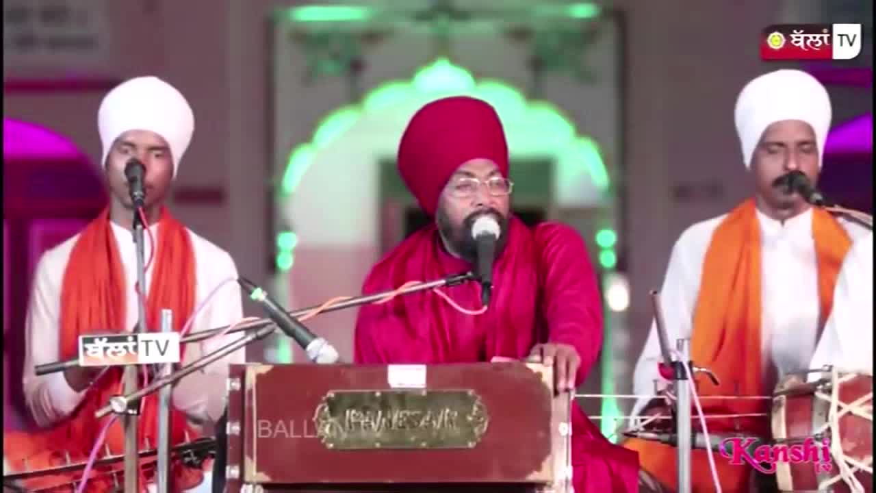 Three men in turbans and orange scarves stand behind microphones, singing. The man in the middle, wearing a red turban, plays a harmonium as Kanshi TV broadcasts the performance.
