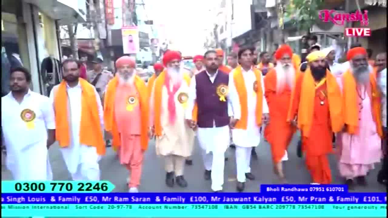 A procession of men in white and orange robes, many with turbans, walks down the street. The Kanshi TV logo is visible in the top right corner, and a phone number and donation information are displayed at the bottom of the screen.
