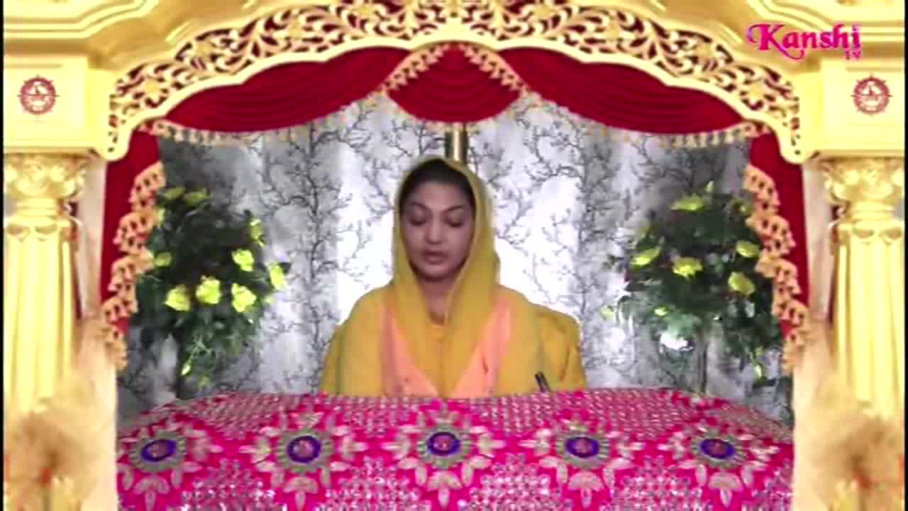 A woman in a yellow head covering speaks from behind a table decorated with pink and gold embroidery. The Kanshi TV logo is visible in the upper right corner.
