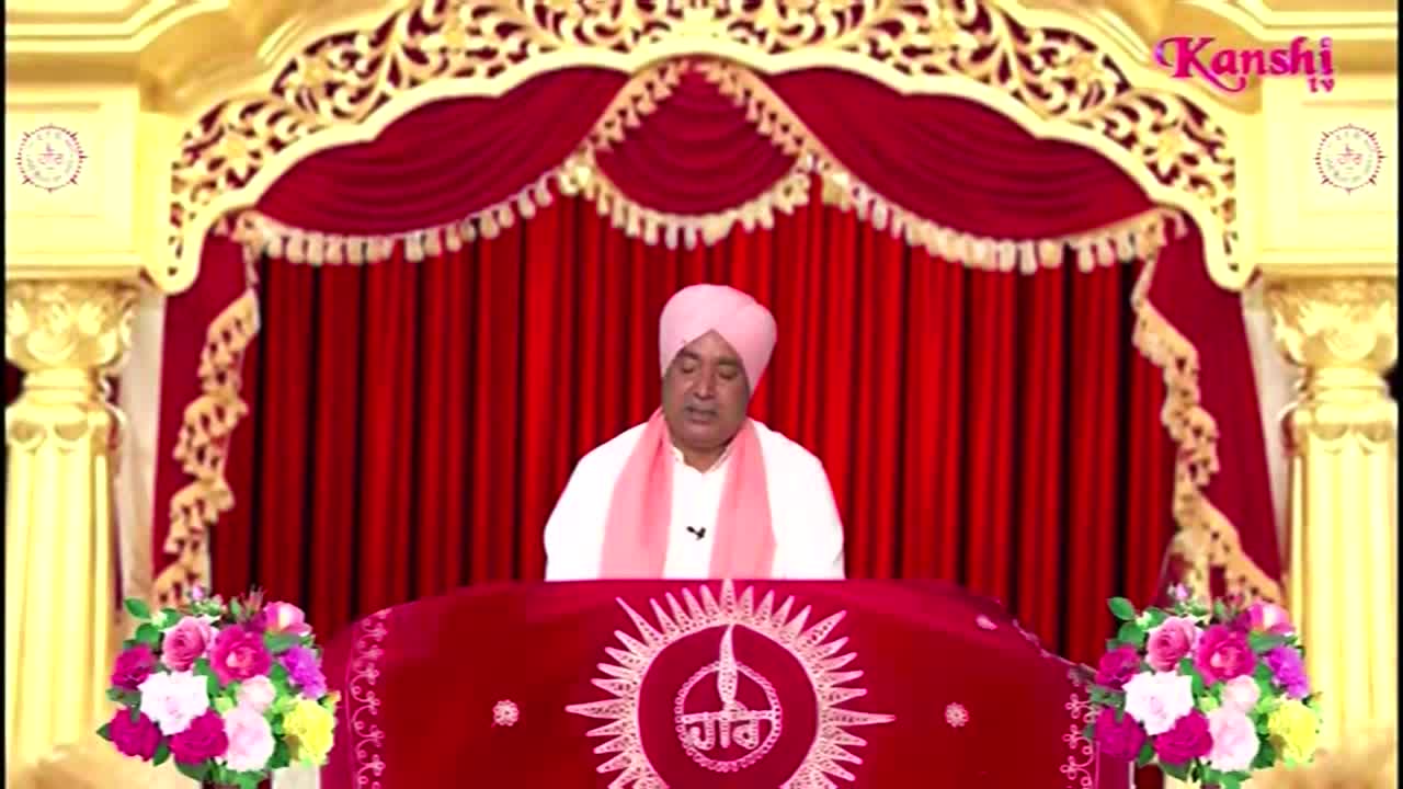 A man in a pink turban and scarf sits behind a red-covered podium, speaking into a microphone. The Kanshi TV logo is visible in the top right corner.

