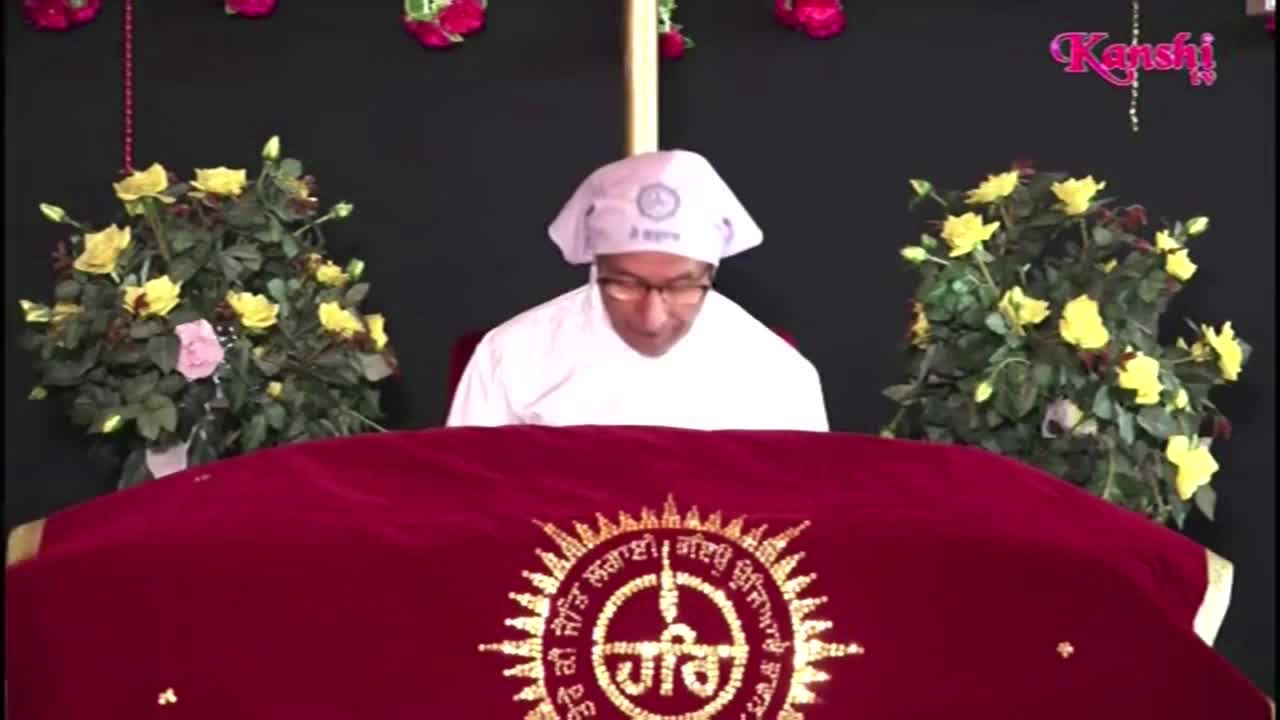 A man in a white turban and glasses reads from behind a red velvet-covered table. The Kanshi TV logo is visible in the upper right corner of the screen.
