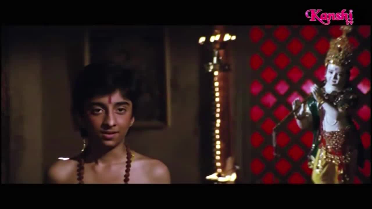 A young man, adorned with a necklace, looks directly at the camera, a small red mark on his forehead. To his right, a statue of a deity stands before a red, diamond-patterned wall, all under the Kanshi TV logo.
