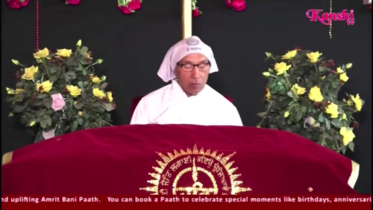 A man in white reads from behind a red cloth-covered table, flanked by floral arrangements. The Kanshi TV logo is visible in the upper right corner.
