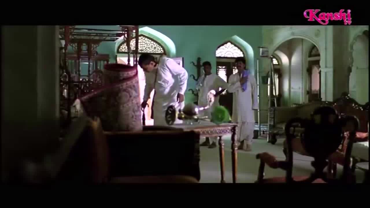 A man in white leans over a table, seemingly inspecting something on it, while two others watch from behind. The scene is set in a large, ornate room, likely part of a film on Kanshi TV.

