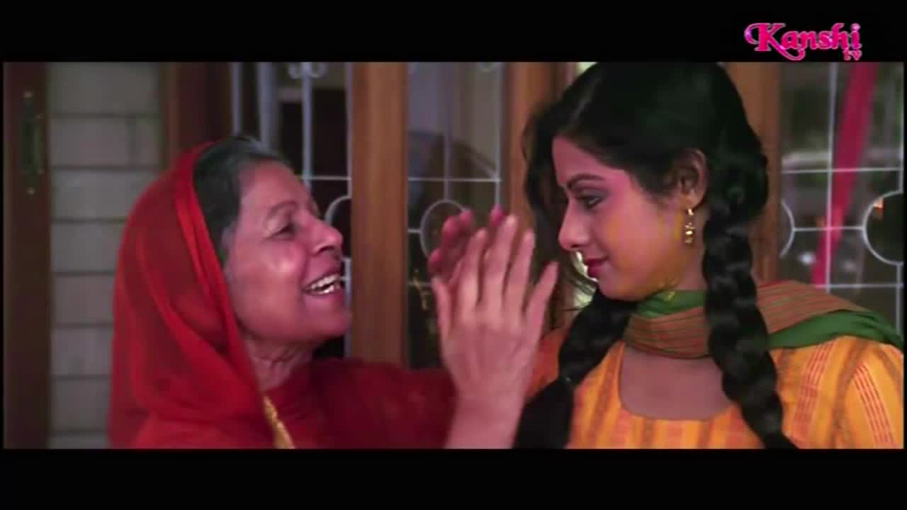 An older woman in a red headscarf is smiling, her hands raised towards the younger woman's face. The younger woman, with her hair in braids, seems to be smiling as well, her face lightly dusted with color.
