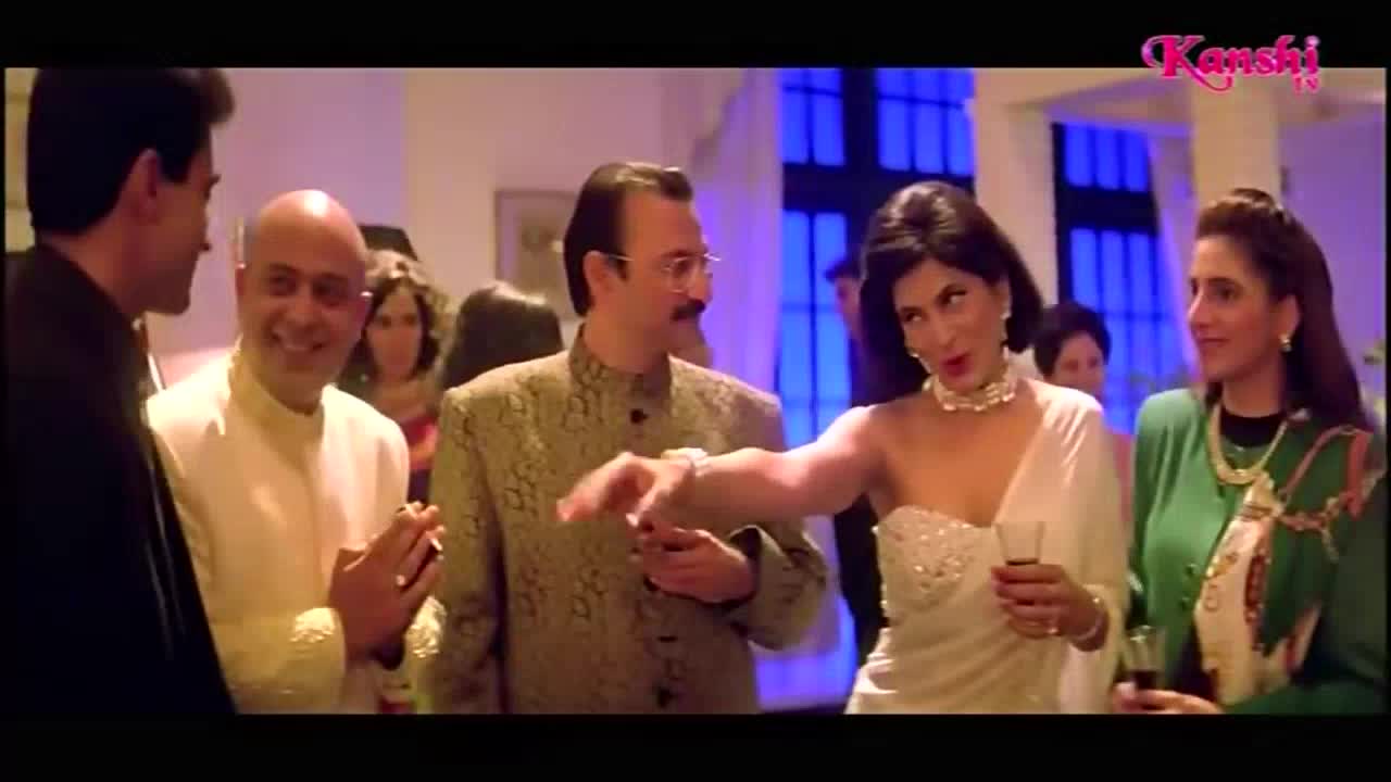 At a party, a woman in a white dress gestures emphatically while holding a glass. The Kanshi TV logo sits in the top right, suggesting a broadcast from the United Kingdom.
