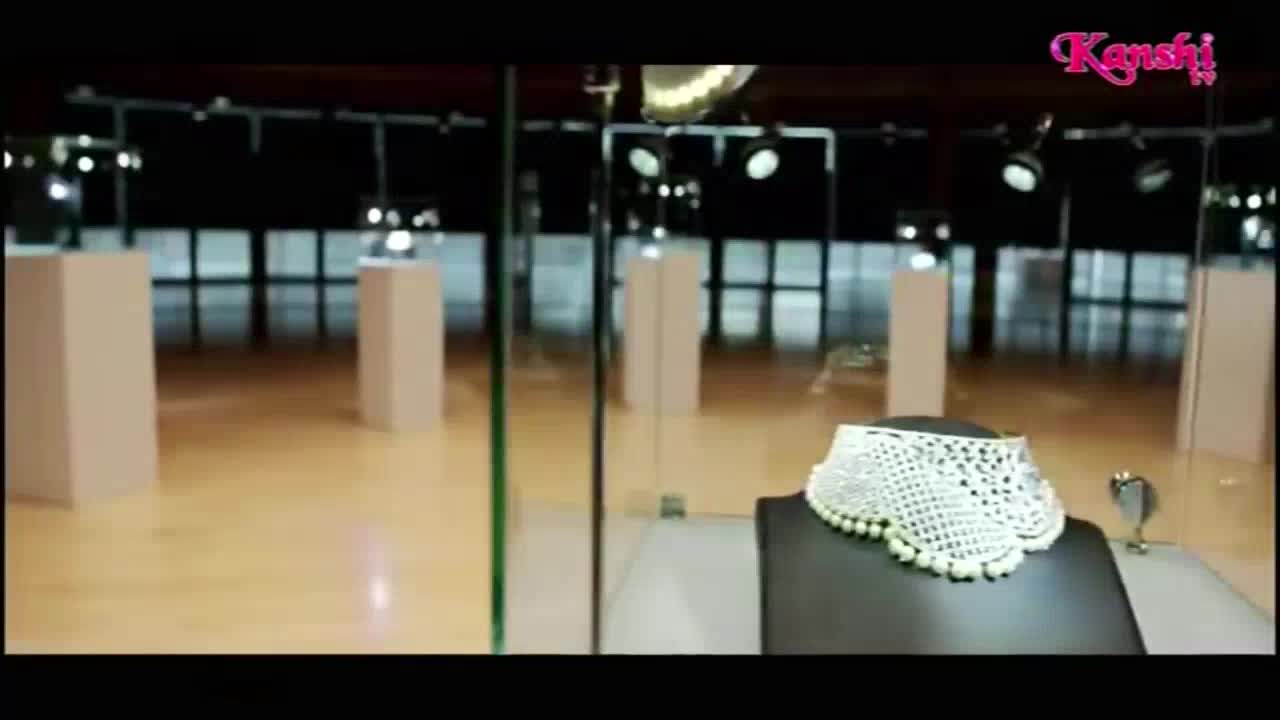 A pearly, lace-like necklace sits on a black stand, lit by overhead spotlights. The Kanshi TV logo floats in the corner, above a display of jewelry in a glass case.
